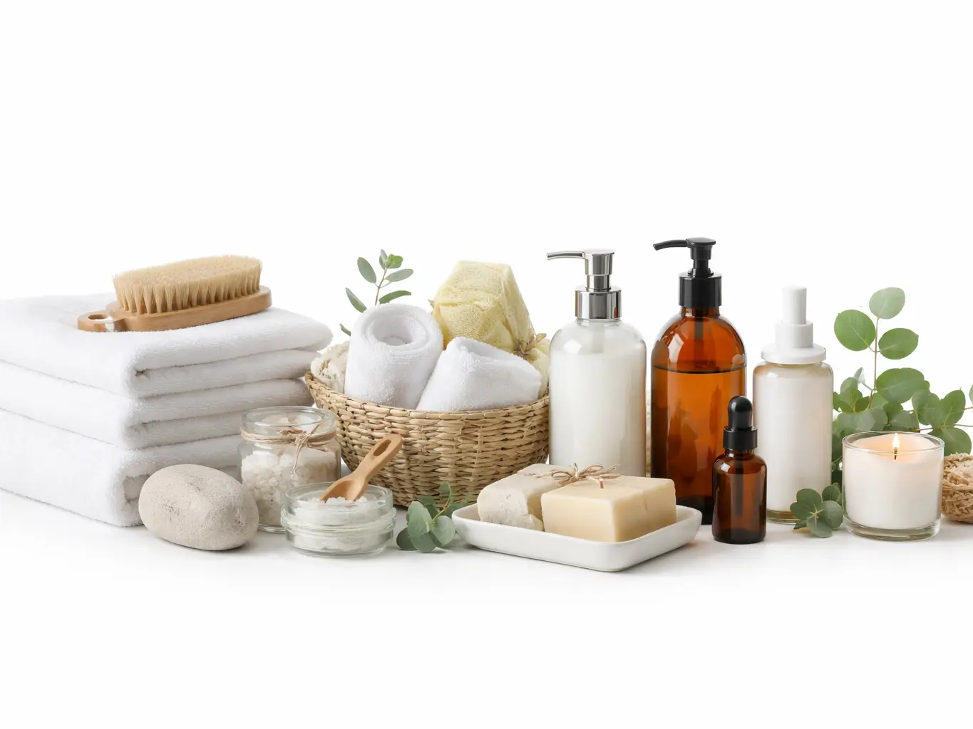 An assortment of luxury spa and bath amenities including stacked white towels, a wooden body brush, bar soaps, bath salts in glass jars, and various amber and white pump bottles with eucalyptus leaves.