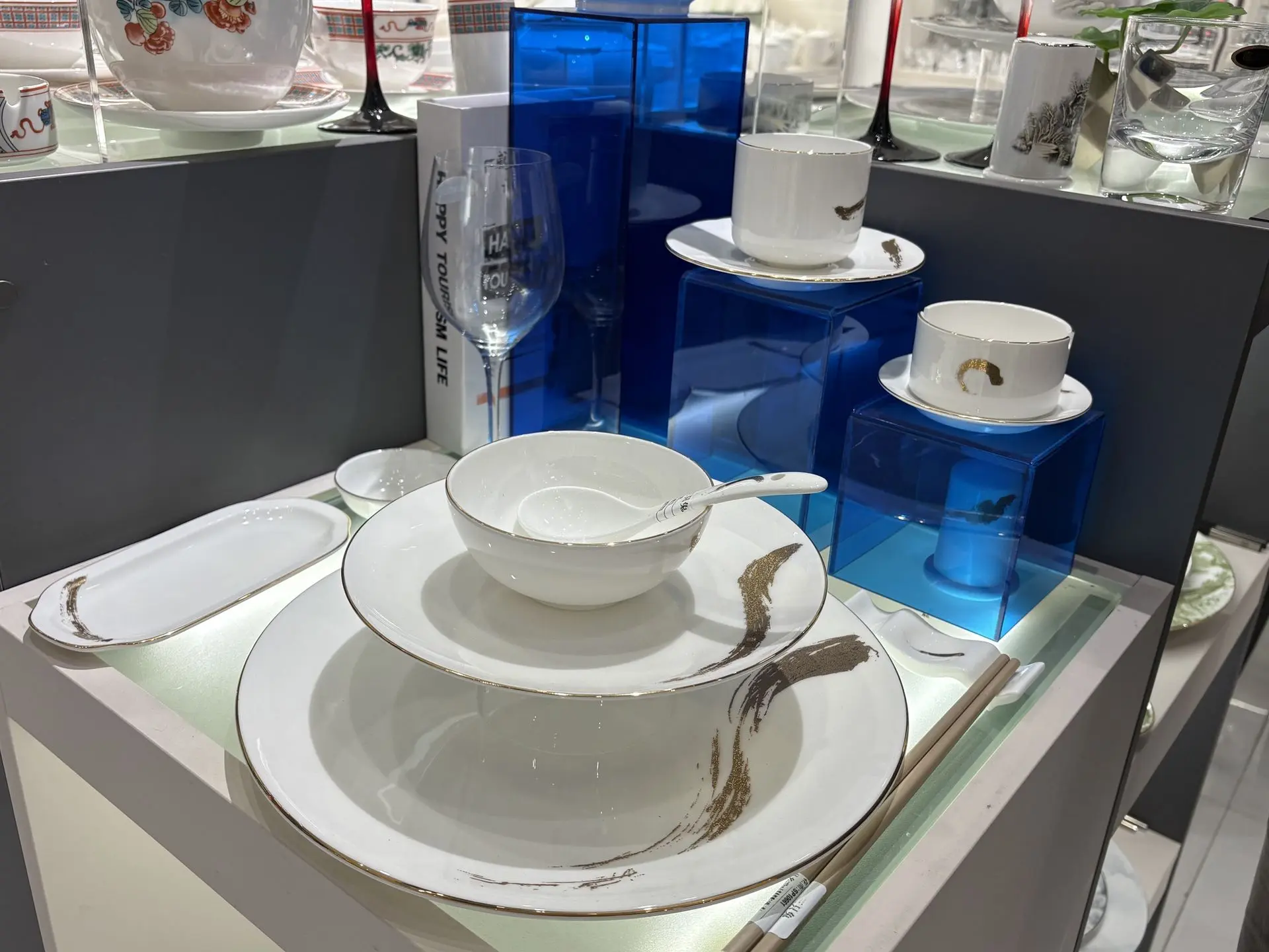 Modern white bone china dinnerware set with gold brushstroke accents, including plates, bowls, and tea cups on a display shelf.