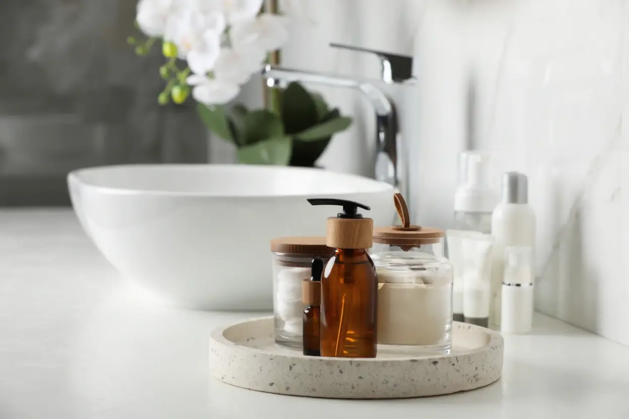 Modern bathroom vanity with a white vessel sink, featuring a stone tray holding amber glass skincare bottles with wooden caps and white cotton pads.