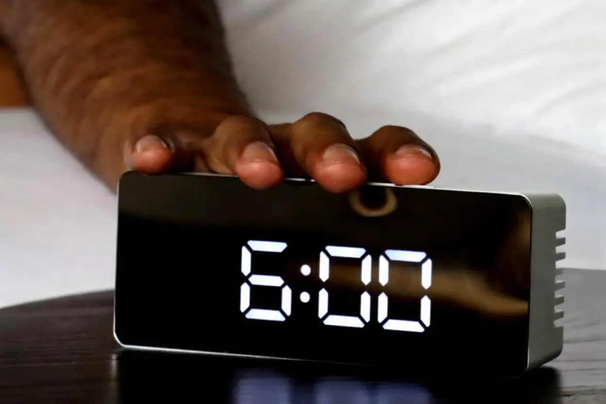 A hand reaching out to press the snooze button on a digital mirror alarm clock displaying 6:00 AM.