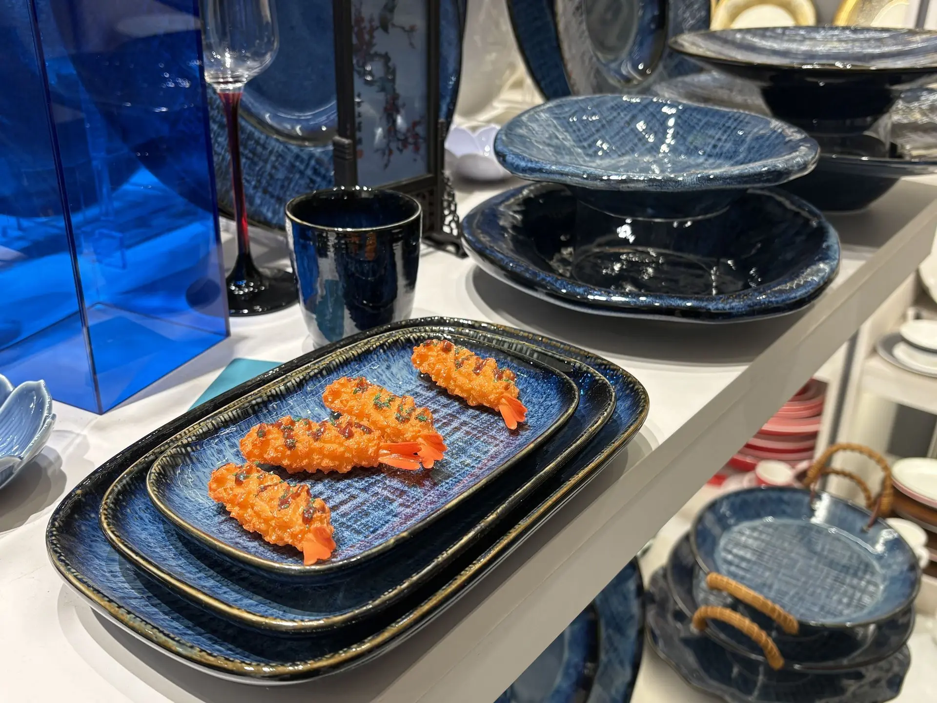 Textured cream and gold Chinese dinnerware set displayed on white retail shelves. A shelf display of cream-colored Chinese crockery featuring a brown sunburst-textured pattern on the rims, including square plates, large oval platters, and matching bowls.