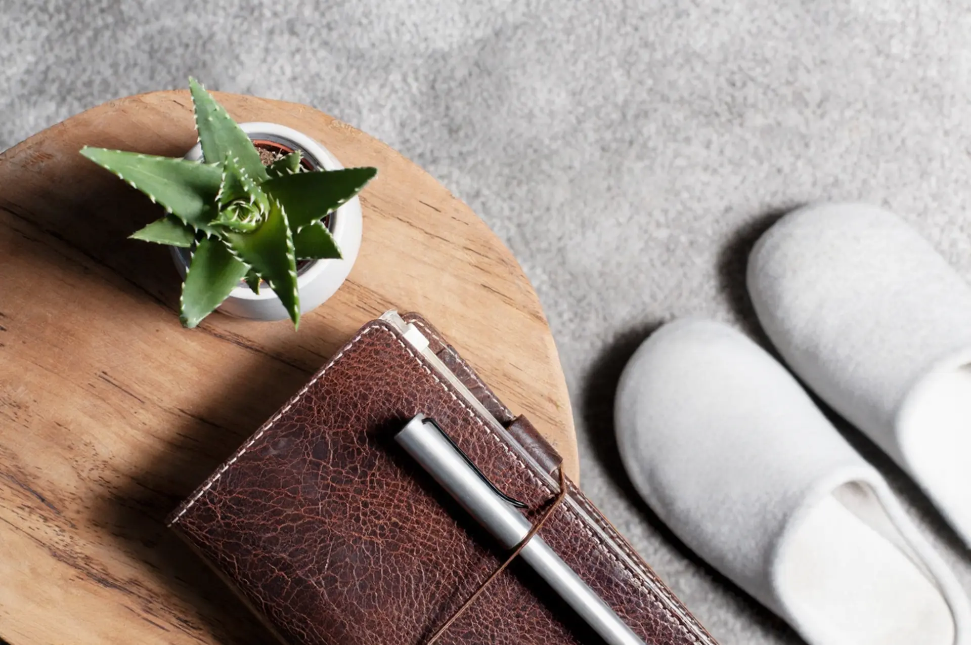 Top-down view of a leather notebook, pen, and succulent on a wooden table next to white hotel slippers in China.