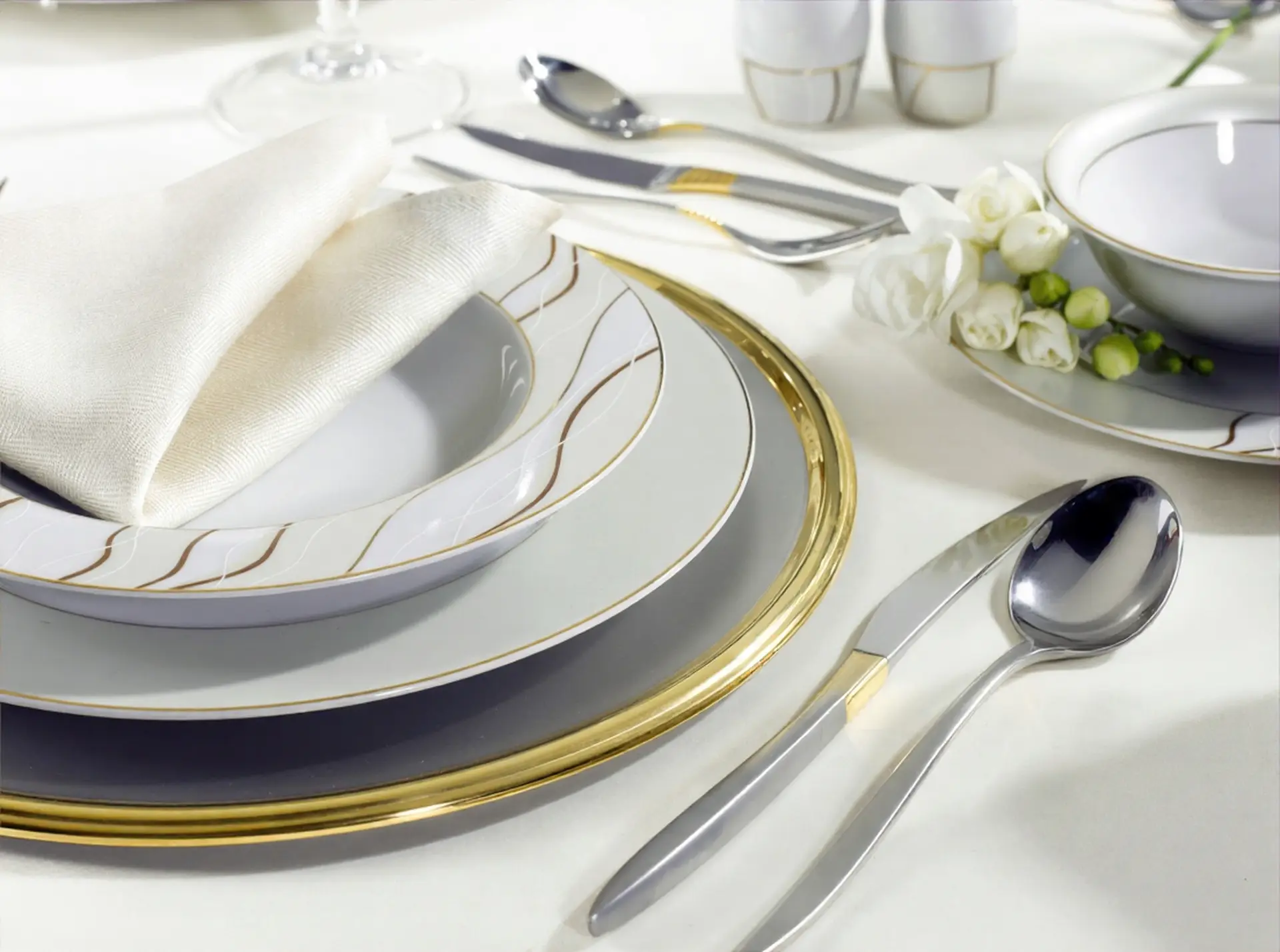 Elegant white and gold fine china dinnerware set with polished silver cutlery and a cream napkin on a white tablecloth.