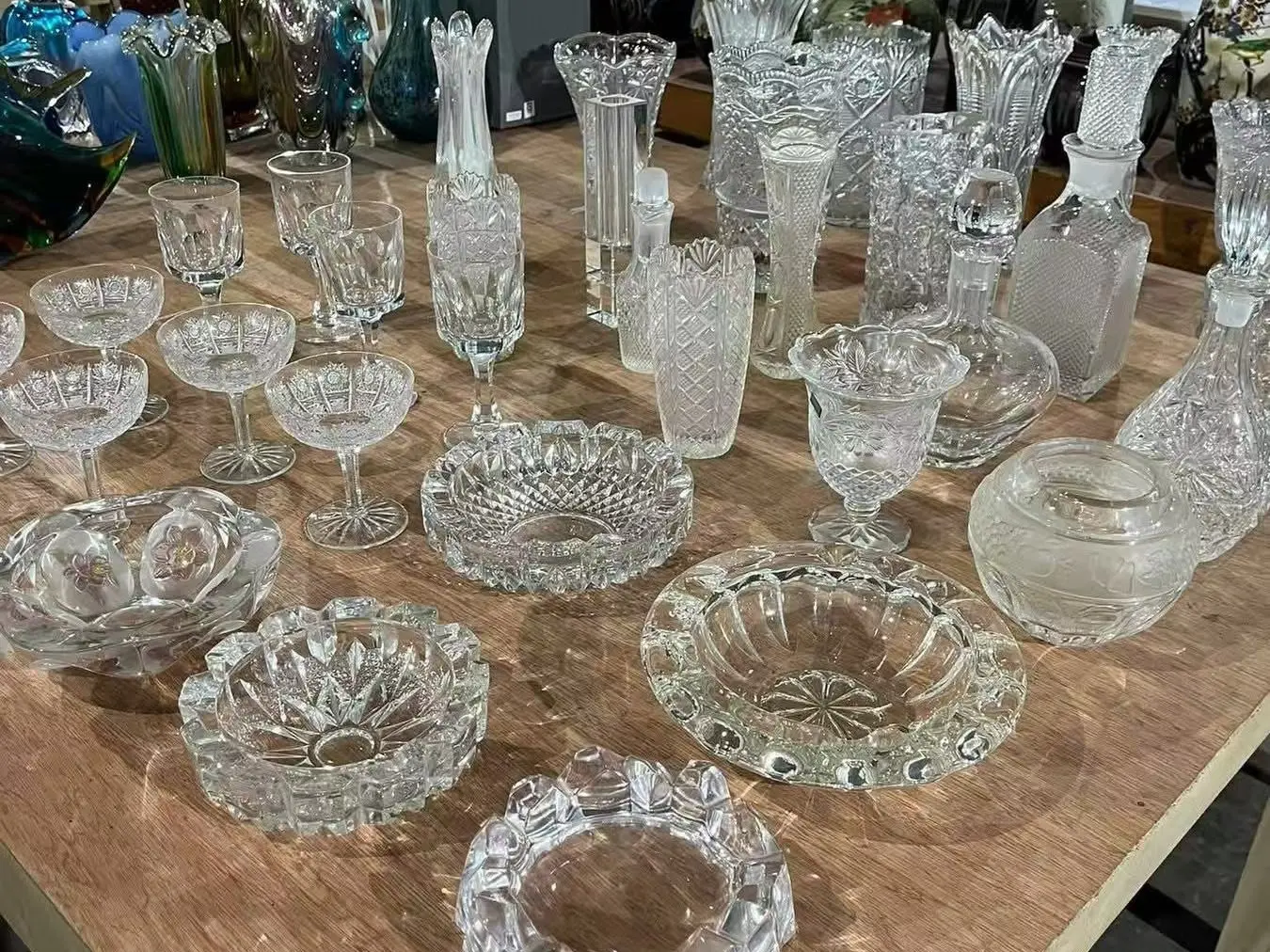 A large collection of assorted crystal glassware, decanters, and ashtrays displayed on a wooden table, featuring various patterns of OS&E from China.
