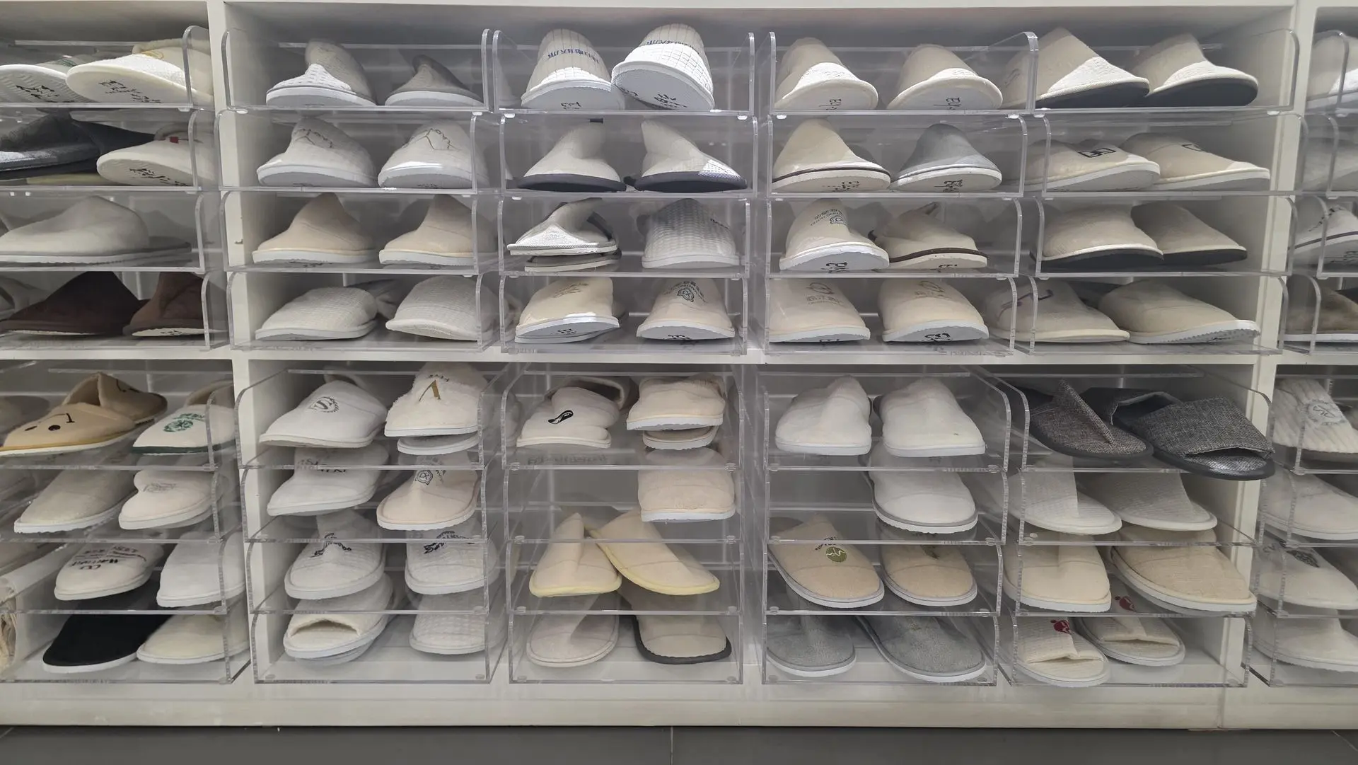 High-Quality China OS&E Slippers Display in Hotel Storage A wall-mounted shelving unit featuring clear acrylic dividers filled with various styles of white, beige, and grey hotel slippers, showcasing luxury China OS&E (Operating Supplies and Equipment) for hospitality.