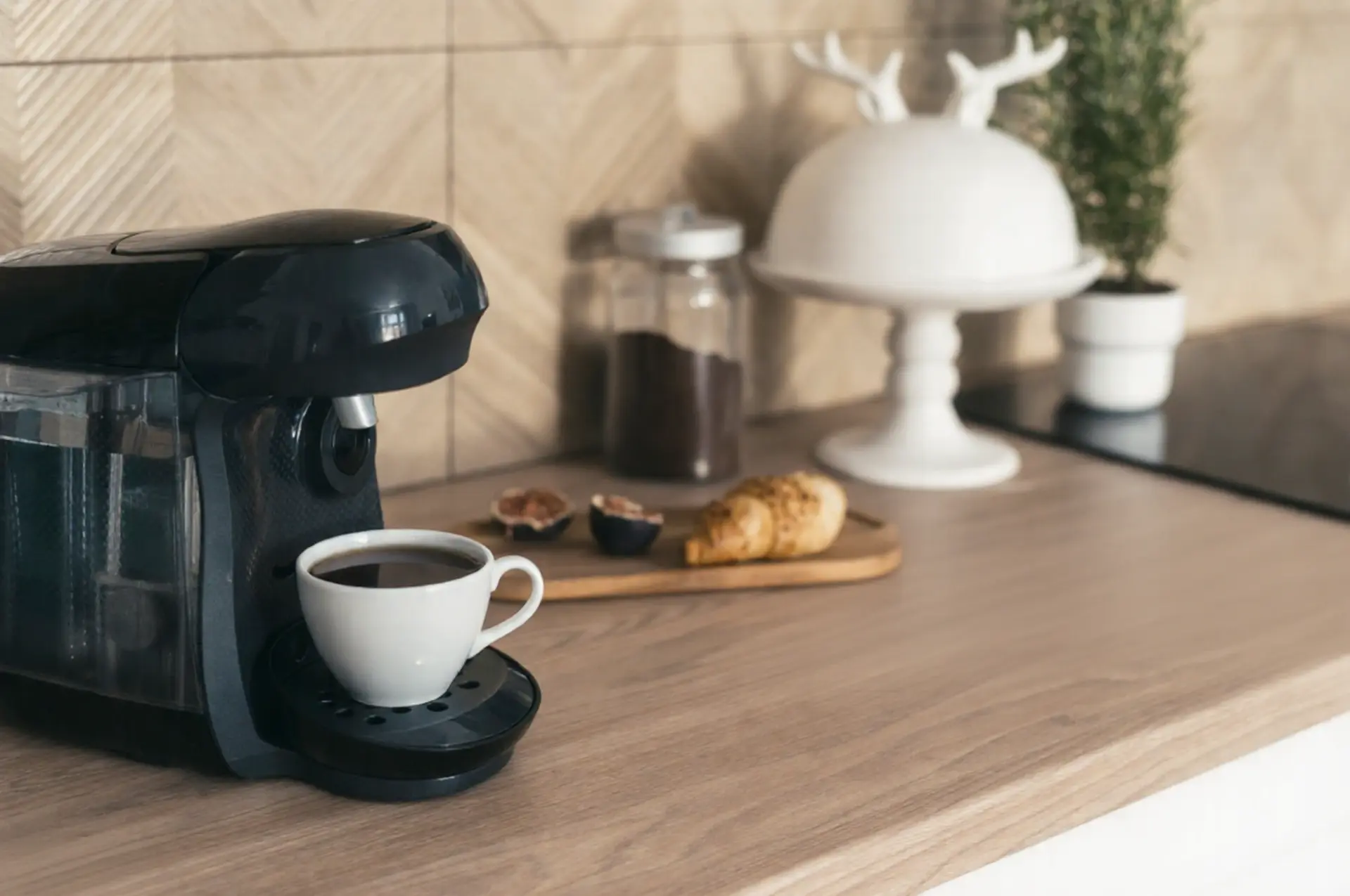 A modern black espresso machine brewing a fresh cup of coffee on a light wooden countertop, accompanied by a croissant and fruit on a wooden board in a stylish kitchen.
