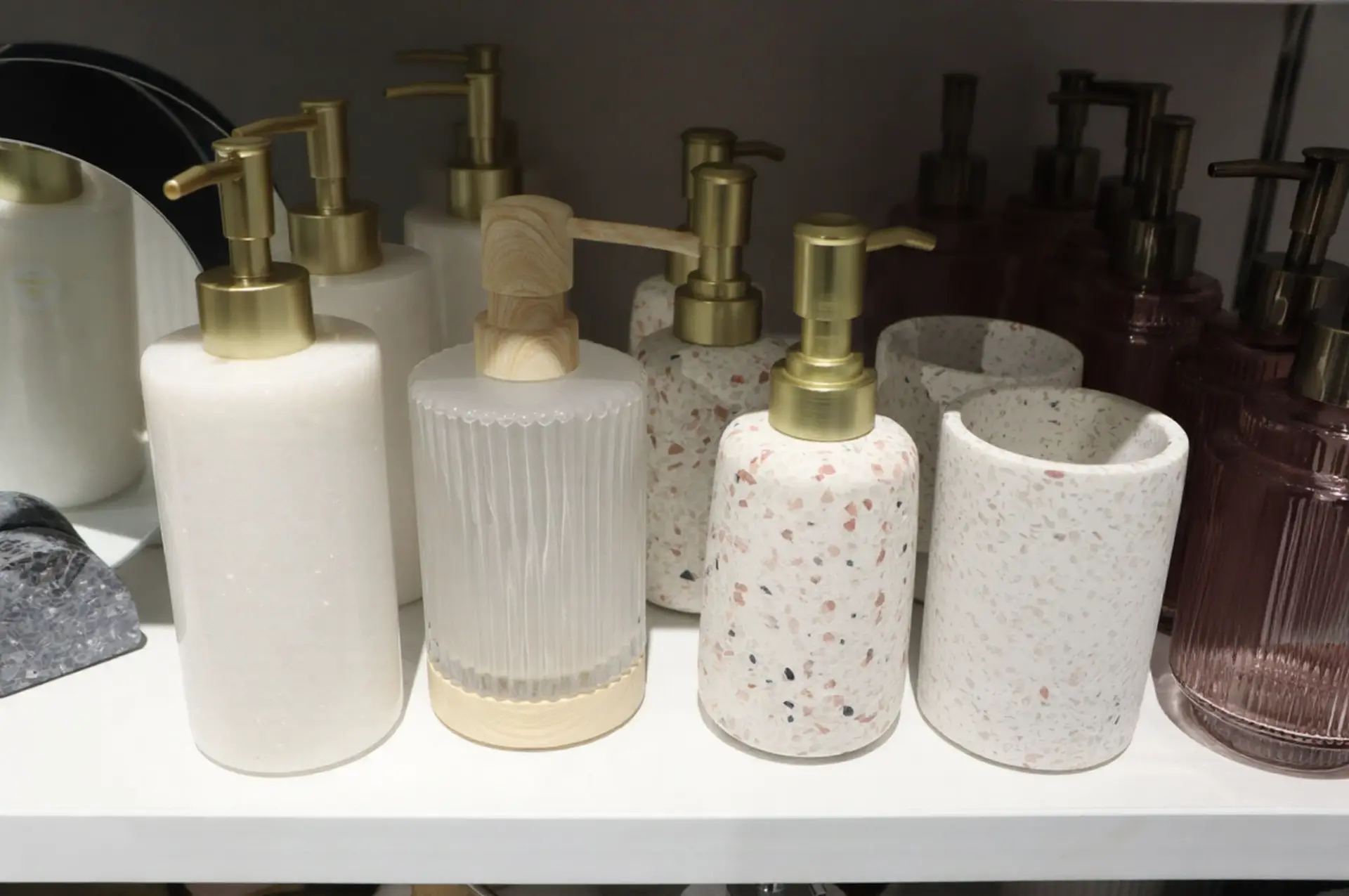 A collection of elegant bathroom accessories featuring soap dispensers in marble, frosted glass, and terrazzo patterns with gold and wood pump accents.