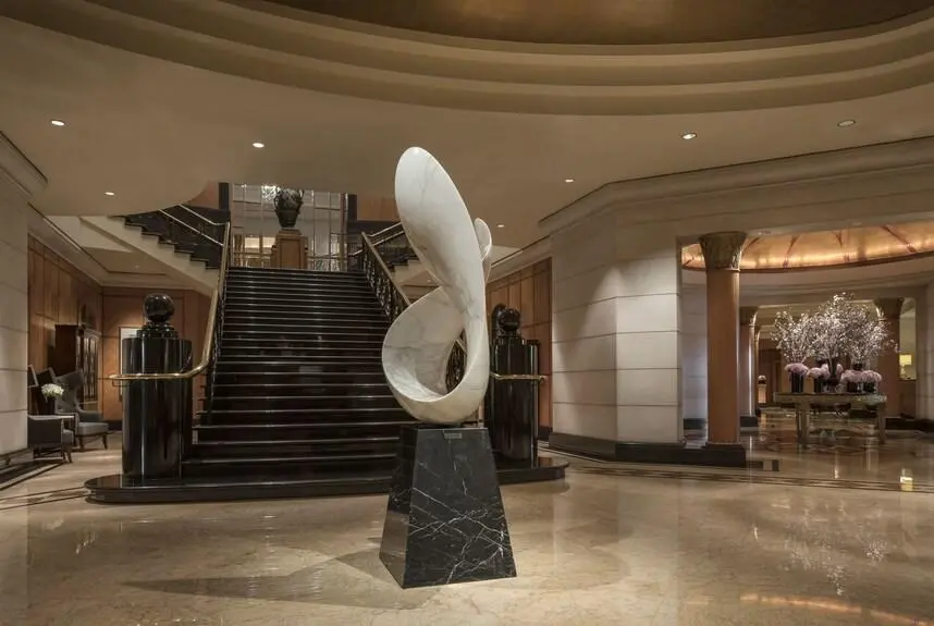 An elegant white marble abstract loop sculpture on a black pedestal, featuring Custom Sculptures from China in a classic luxury hotel lobby with a grand staircase.