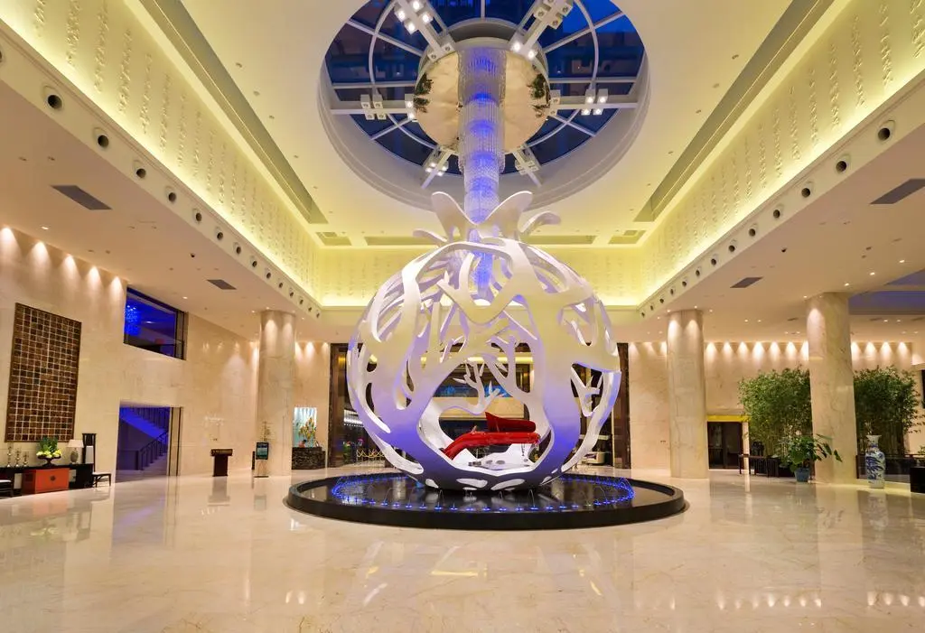 A massive white spherical lattice sculpture housing a red piano, representing Custom Sculptures from China, situated in a grand marble hotel lobby under a skylight