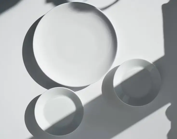 Top-down view of minimalist white crockery including a dinner plate and two bowls arranged on a surface with dramatic shadows.
