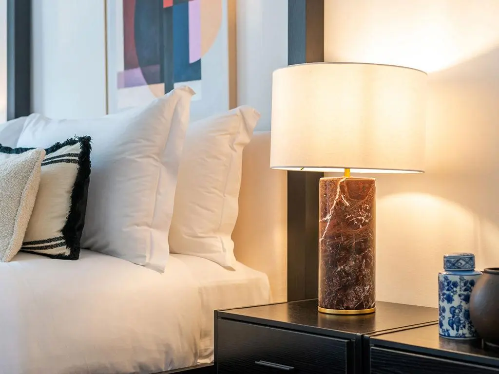 Luxury guest room decor featuring a cylindrical brown marble table lamp on a dark wood nightstand next to a bed dressed with crisp white linens, decorative throw pillows, and abstract wall art Luxury Guest Room Decor with Marble Table Lamp and Abstract Art