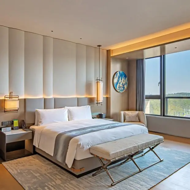 Modern Hotel Bedroom Featuring a Neutral Chinese Headboard and Panel Wall Modern hotel bedroom with a neutral upholstered Chinese headboard, vertical panel wall lighting, a large bed with crisp white linens, and a bench at the foot of the bed.